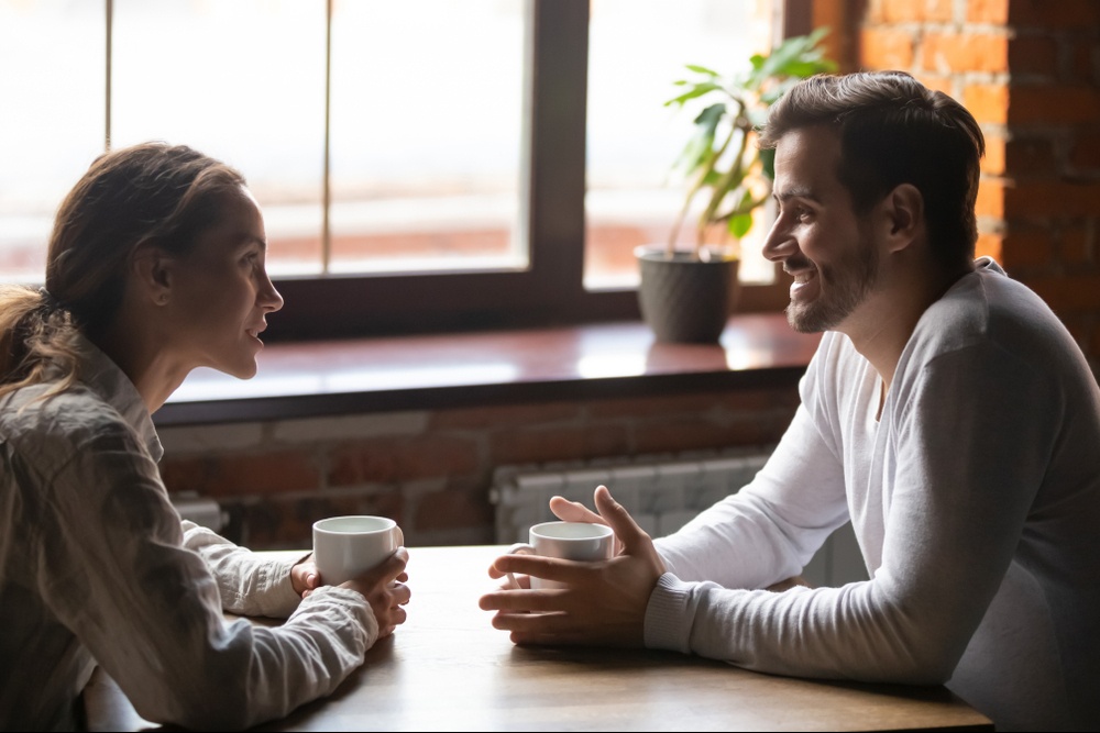A couple holding coffee cups smile while sitting across a table from one another. This could symbolize the benefits on relationships from working with a trauma therapist in Delray Beach, FL. Search for a trauma treatment in Palm Beach County, FL by contacting a therapist in Palm Beach County, FL today.