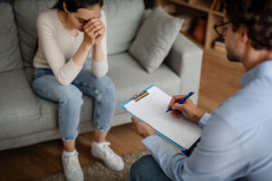 A woman bows her head while sitting across from a person with a clipboard, taking notes. Learn how a trauma therapist in Delray Beach, FL can offer support in addressing bottled feelings. Search for trauma treatment in Palm Beach County, FL to learn more about the benefits of trauma therapy Delray Beach. 