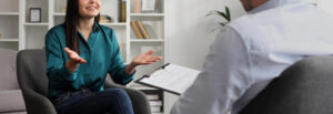 A woman smiles while gesturing with her hands and sitting across from a person with a clipboard. This could represent the support an addiction therapist in Palm Beach County, FL can offer. Learn more about addiction counseling in Miami, FL, and other types of addiction in Palm Beach, FL today. 